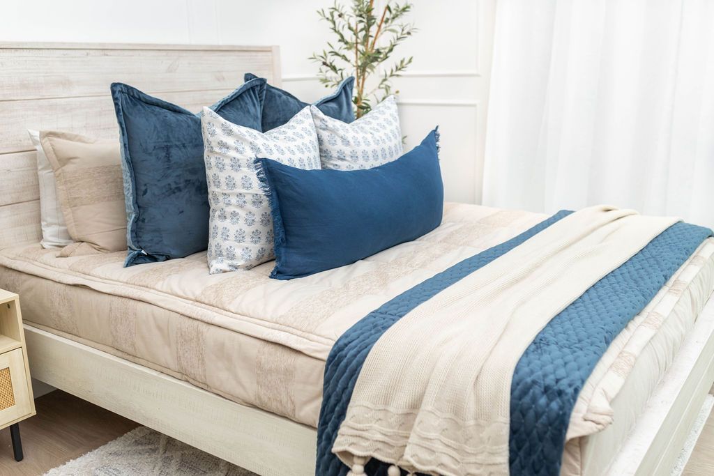 A brightly lit bedroom with a bed made with tan zipper bedding and an assortment of neutral and blue velvet throw pillows and blankets.