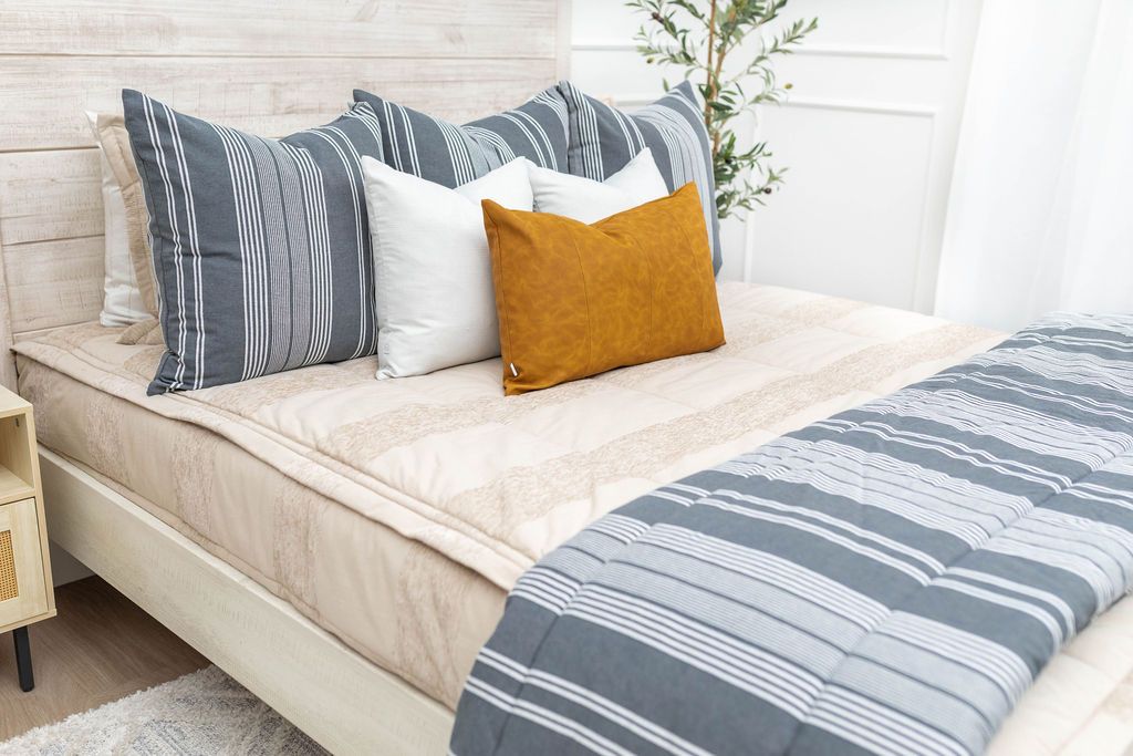 A brightly lit bedroom with a bed made with tan faded stripes zipper bedding and an assortment of neutral and gray striped pillows and blanket.