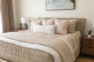 A brightly lit bedroom with a bed made with tan faded stripes zipper bedding and an assortment of neutral and light pink pillows and blankets.