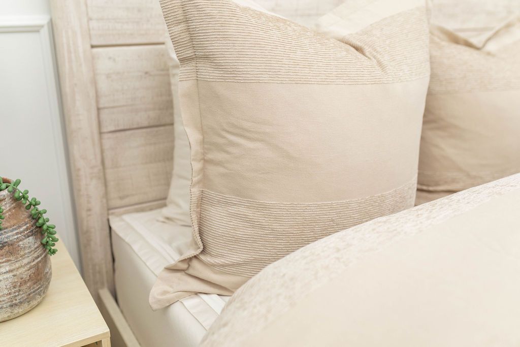 A close up view of a bed made with white zipper sheets and a tan zipper duvet folded partially down the bed and adorned with neutral large matching tan pillows.