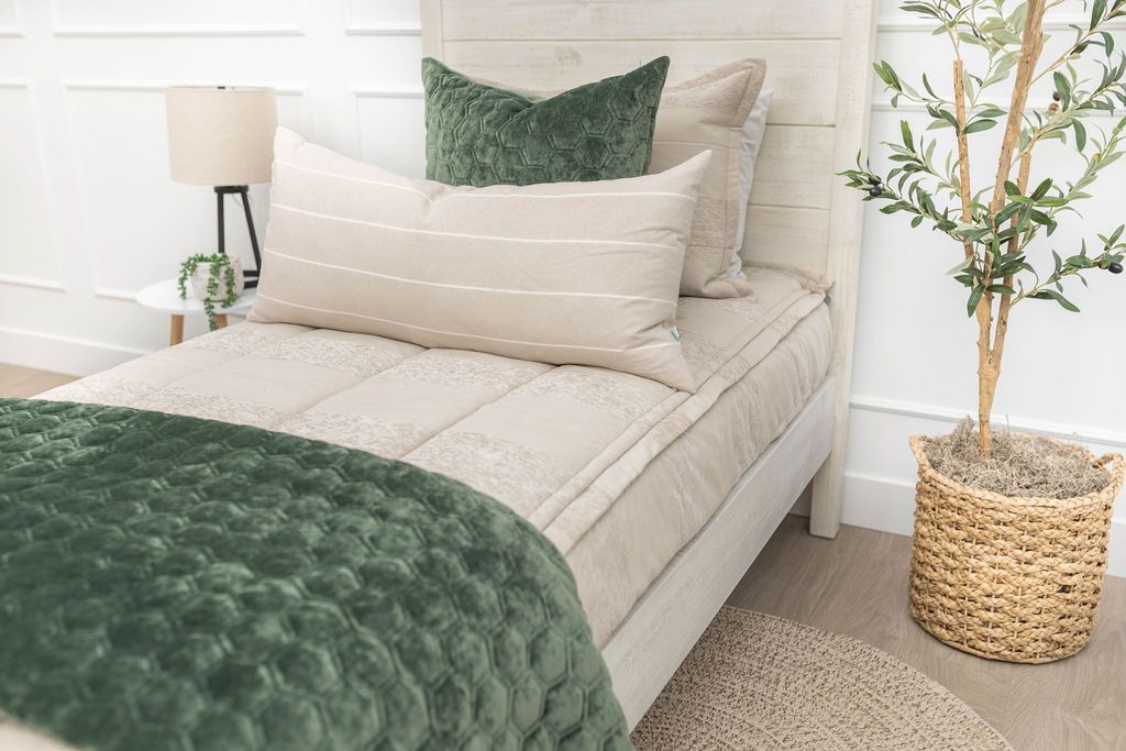 A brightly lit bedroom with a twin bed made with tan faded stripes zipper bedding and an assortment of neutral and green velvet throw pillows and blanket.