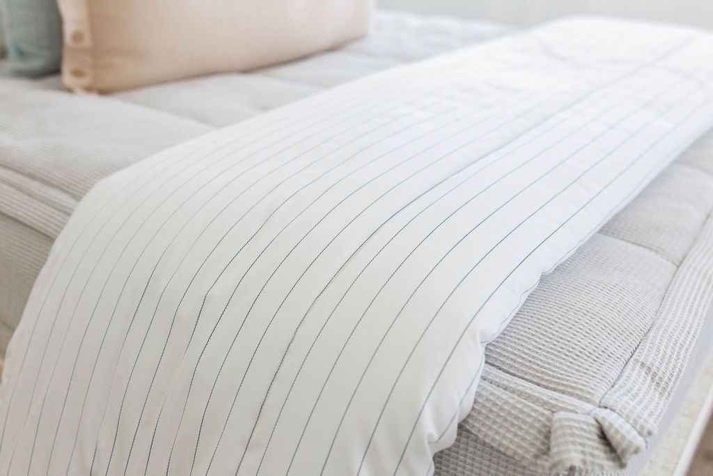A close up of a bed made with light gray zipper bedding and a white blanket with a light thin blue striped pattern at the end of the bed.