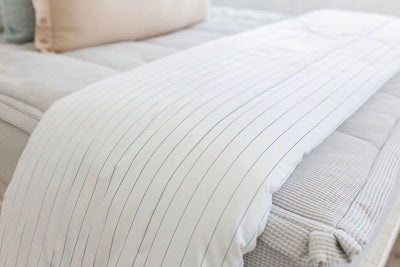A close up of a bed made with light gray zipper bedding and a white blanket with a light thin blue striped pattern at the end of the bed.