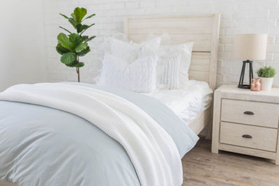 A twin bed made with ruffled white zipper bedding and a light blue zipper duvet folded half way down the bed and adorned with white ruffled pillows and a throw blanket.
