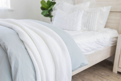 A side view of a bed made with white ruffled zipper bedding and a light blue zipper duvet folded half way down the bed and adorned with white ruffled pillows and matching throw blanket.