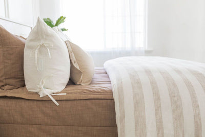A side view of a bed made with brown zipper bedding and a woven ivory and tan striped zipper duvet folded half way down the bed and adorned with neutral pillows.