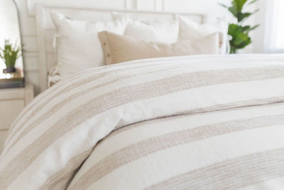 A close up view of a bed made with a woven ivory and tan striped zipper duvet folded half way down the bed and adorned with neutral pillows.