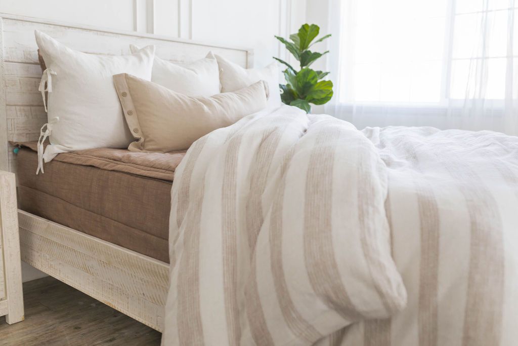 A brightly lit bedroom with a bed made with brown zipper bedding and a woven ivory and tan striped zipper duvet partially folded half way down the bed and adorned with neutral pillows.
