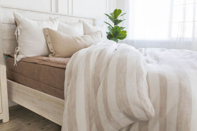 A brightly lit bedroom with a bed made with brown zipper bedding and a woven ivory and tan striped zipper duvet partially folded half way down the bed and adorned with neutral pillows.