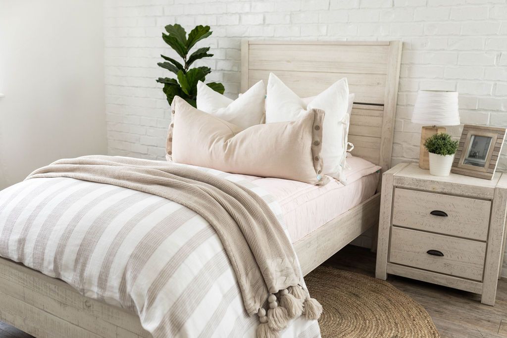 A brightly lit bedroom with a bed made with  light pink zipper bedding and a woven ivory and tan striped zipper duvet folded half way down the bed and adorned with neutral pillows and blanket.