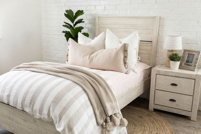 A brightly lit bedroom with a bed made with  light pink zipper bedding and a woven ivory and tan striped zipper duvet folded half way down the bed and adorned with neutral pillows and blanket.