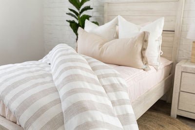 A brightly lit bedroom with a twin bed made with  light pink zipper bedding and a woven ivory and tan striped zipper duvet folded partially half way down the bed and adorned with neutral pillows.