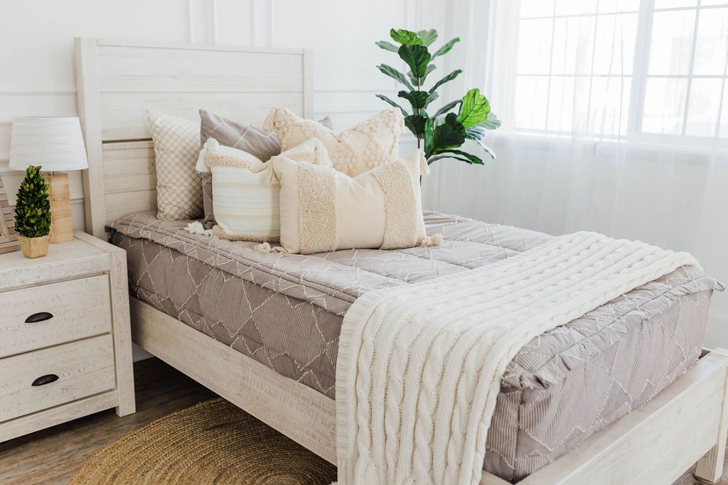 twin bed with Taupe bedding with textured zig zag design dark creamy textured euro, a cream and tan woven textured pillow and a textured dark creamy lumbar with tassels with an off white braided throw with pom poms along the edge