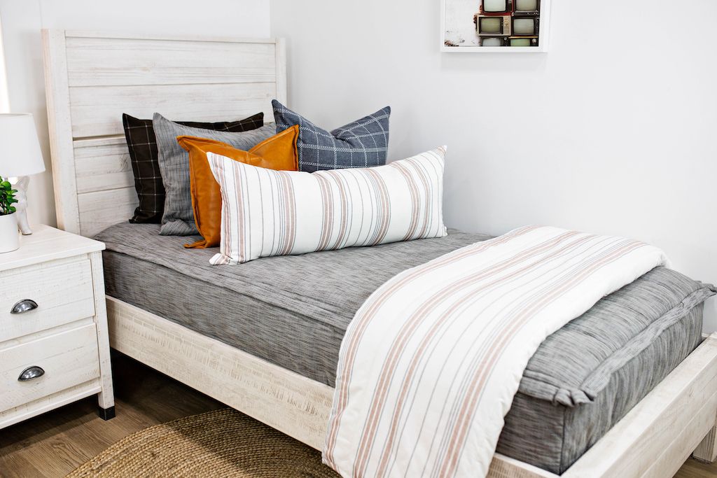White twin size bed with gray and brown woven bedding, blue and gray plaid pillows, cream and cognac striped lumbar pillow and a cream and cognac striped blanket.
