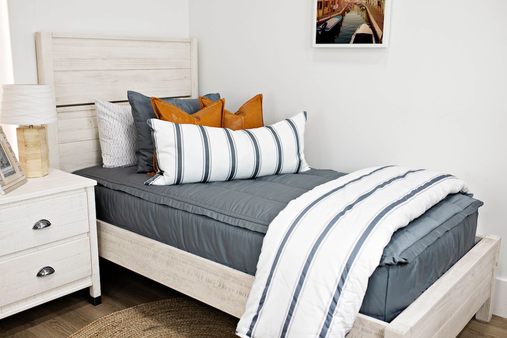 White twin size bed with gray bedding, medium brown leather pillows, a white and gray striped lumbar pillow and a white and gray striped blanket.  