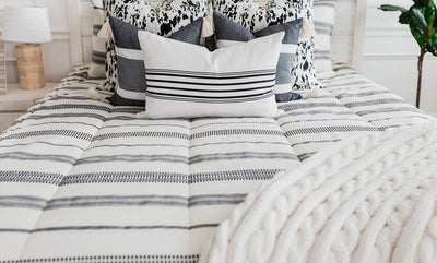 Queen bed with cream and black woven striped bedding, a white and black euro pillow, a medium black and cream textured pillow, a cream and black striped lumbar pillow, a cream cable-knit throw at the foot of the bed.