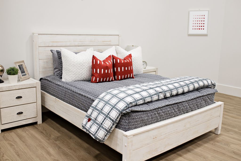 queen bed with Gray bedding with textured diamond pattern and white faux fur textured euros, red and white dashed pillows with white and gray plaid blanket with an embroidered red heart in the corner