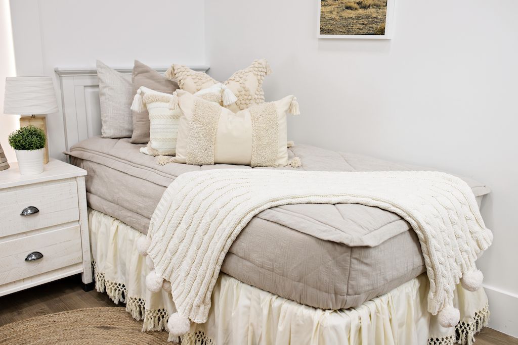 twin bed with tan textured bedding and dark creamy textured euro, a cream and tan woven textured pillow and a textured dark creamy lumbar with tassels with an off white braided throw with pom poms along the edge with cream boho bedskirt 