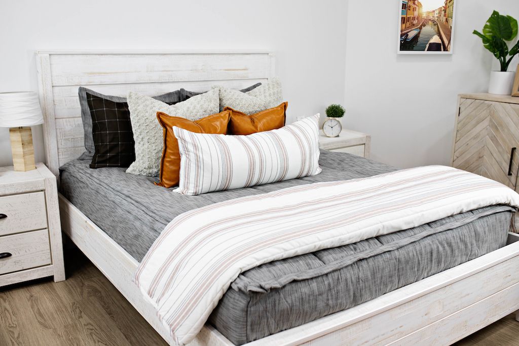 White queen bed with gray and brown woven bedding, cream textured pillows, brown leather pillows, a cream and cognac striped lumbar pillow and a cream and cognac blanket.