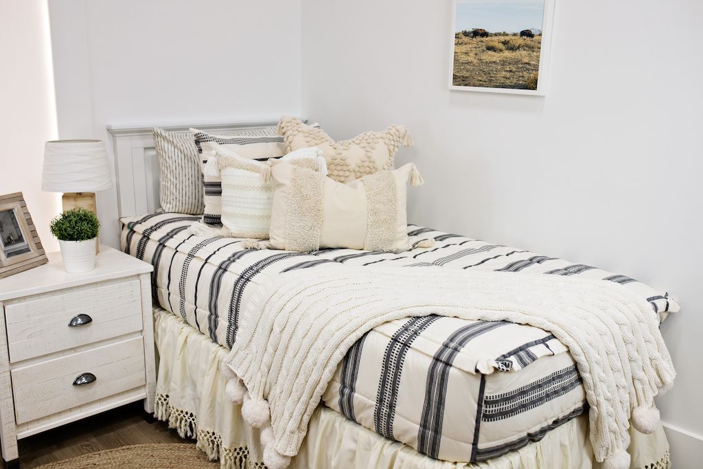 White twin bed with white and black woven bedding, a cream textured euro pillow, a medium cream textured pillow, a cream textured lumbar pillow and a cream throw.  