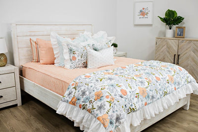 White queen bed with peach textured bedding, white and blue striped pillows, multicolored floral pillows, a gray and white lumbar pillow and a multicolored floral blanket.