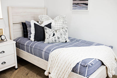 twin bed with blue striped bedding and cream and black grid euro with ruffle along the edge, charcoal striped pillow with white ruffle along the edge, cream lumbar with charcoal paisley print, cream knitted chenille blanket with pom poms