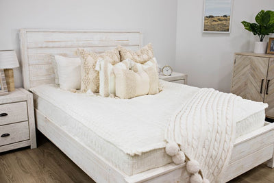 queen bed with off white textured bedding and dark creamy textured euro, a cream and tan woven textured pillow and a textured dark creamy lumbar with tassels with a cream knitted chenille throw pom poms along the edge