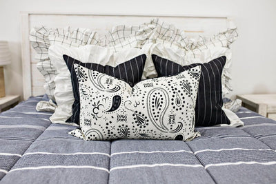 two cream and black grid euro with ruffle along the edge, two charcoal striped pillow with white ruffle along the edge, cream lumbar with charcoal paisley print, cream knitted chenille blanket with pom poms