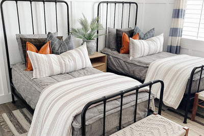 two black metal twin bed frames with Brown and gray woven textured bedding with blue grid euro, faux leather pillow, cream striped XL lumbar and cream striped blanket at the foot of the bed