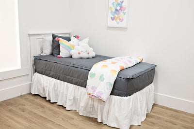 White twin size bed with gray bedding, a medium white and rainbow heart pillow, a medium white rainbow pillow, a white cloud shaped pillow, a white bed skirt and a white and rainbow heart blanket at the foot of the bed.