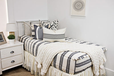 White twin size bed with cream and black woven striped bedding, a white and black euro pillow, a medium black and cream textured pillow, a cream and black striped lumbar pillow, a cream bed skirt with tassels and a cream cable-knit throw at the foot of the bed. 