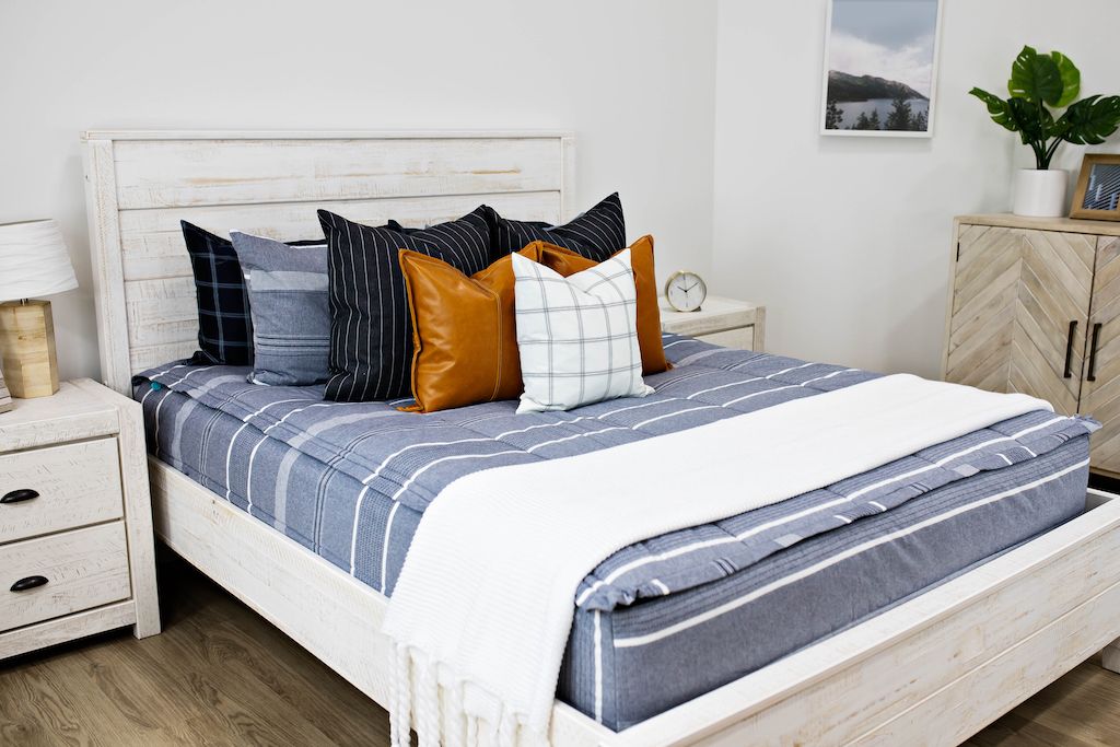 queen bed with blue and navy striped bedding and deep navy striped euros, faux leather pillows, white and black grid pillow, and white textured blanket with braided tassels