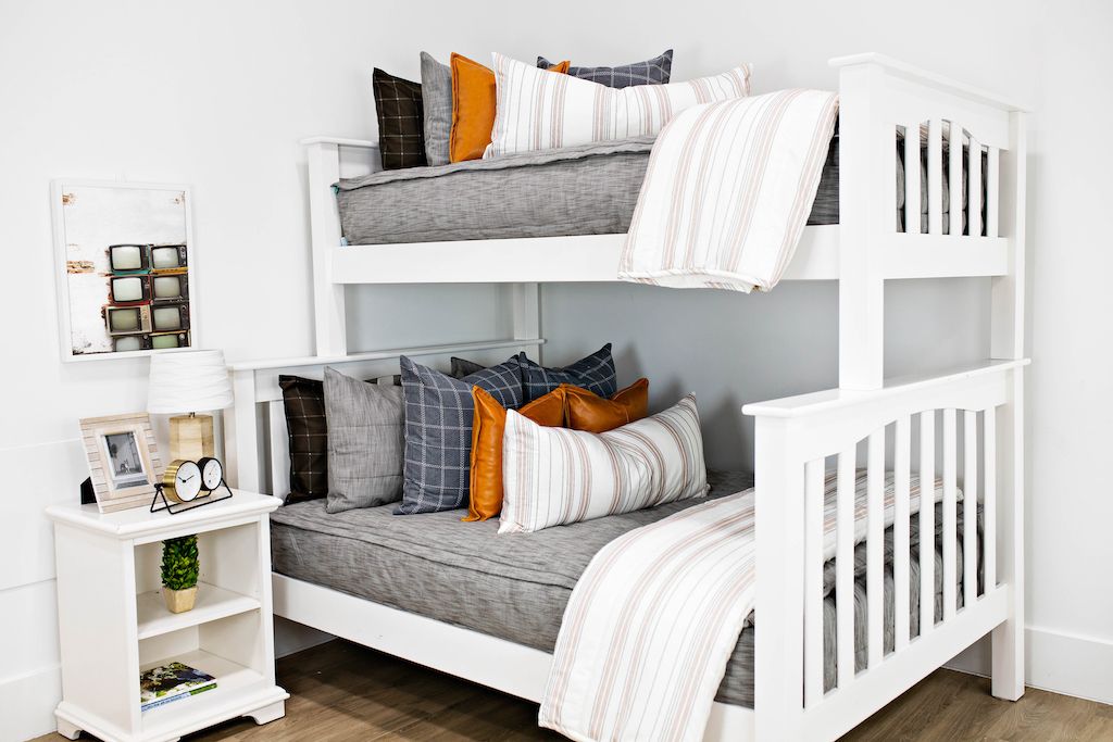 White bunk beds with gray and brown woven bedding, blue and gray plaid pillows, cream and cognac striped lumbar pillows and cream and cognac striped blankets. 