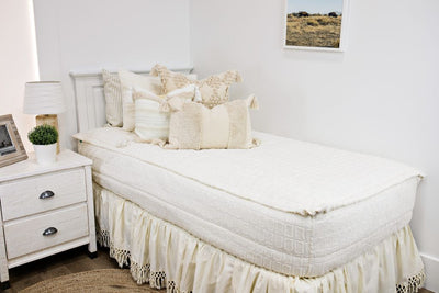 White twin size bed with cream textured bedding, a cream textured euro pillow, a medium cream textured pillow, a cream textured lumbar pillow and a cream bed skirt with tassels.