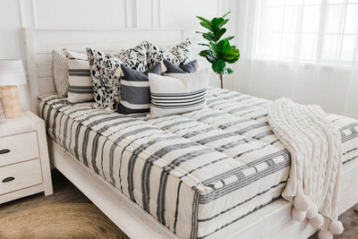 Queen bed with cream and black woven striped bedding, a white and black euro pillow, a medium black and cream textured pillow, a cream and black striped lumbar pillow, a cream cable-knit throw at the foot of the bed.