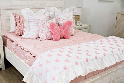 queen bed with Blush pink bedding with pink and white striped euro with ruffle, white and pink butterfly print pillow, pink plush butterfly pillow, and white and pink print blanket with white ruffle along the edge