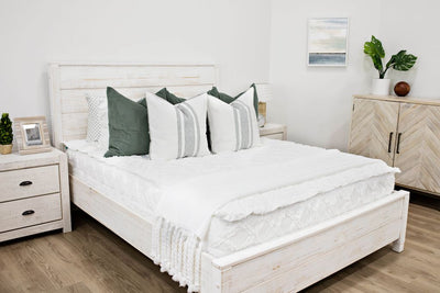White queen bed frame decorated with white textured bedding, White Euro pillows with moss green striping design with moss green stitching along the edge with moss green corduroy euro pillow behind and a textured white throw with braided tassels with horizon artwork on the wall