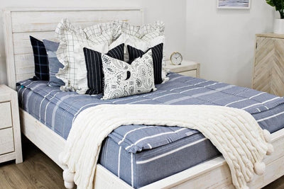 White queen bedframe with blue striped bedding, cream and black striped euros with ruffle along the edge, charcoal striped pillow with white ruffle along the edge, cream lumbar with charcoal paisley print, and cream textured blanket with pom poms