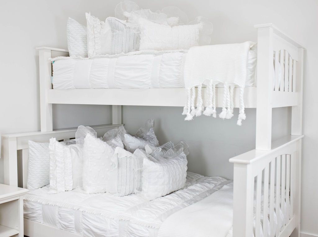 White bunk bed with white textured bedding, white ruffle polka dot euros,  ruffle gray/blue textured pillows, white ruffle textured lumbar, and a textured white throw with braided tassels