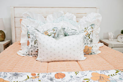Enlarged view of a white bed with peach textured bedding, white and blue striped pillows, multicolored floral pillows and a gray and white lumbar pillow.  