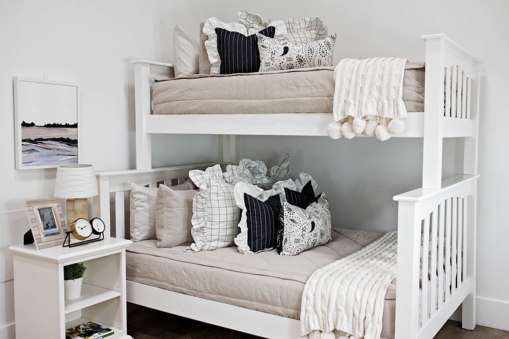 bunk bed with tan textured bedding and cream and black grid euro with ruffle along the edge, charcoal striped pillow with white ruffle along the edge, cream lumbar with charcoal paisley print, cream knitted chenille blanket with pom poms