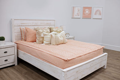 queen bed with peach textured bedding and dark creamy textured euro, a cream and tan woven textured pillow and a textured dark creamy lumbar with tassels