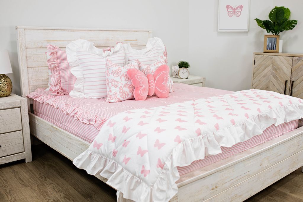White queen bed with pink and white striped ruffled bedding, white and pink striped pillows, white and pink butterfly pillows, a pink butterfly shaped pillow and a white and pink butterfly blanket.  