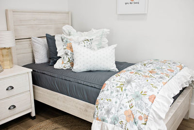 White twin size bed with gray bedding, a white and blue striped pillow, a multicolored floral pillow, a gray and white lumbar pillow and a multicolored floral blanket.  