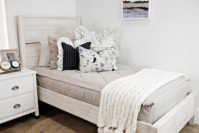 twin bed with tan textured bedding and cream and black grid euro with ruffle along the edge, charcoal striped pillow with white ruffle along the edge, cream lumbar with charcoal paisley print, cream knitted chenille blanket with pom poms