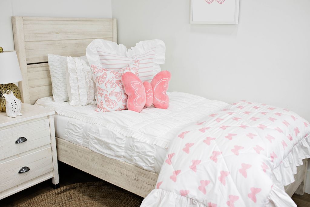 White twin size bed with white textured bedding, a white and pink striped pillow, a white and pink butterfly pillow, a pink butterfly shaped pillow and a white and pink butterfly blanket.  