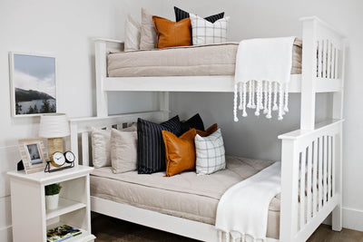 bunk bed with tan textured bedding and deep navy striped euros, faux leather pillows, white and black grid pillow, and white textured blanket with braided tassels