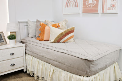White twin size bed with beige textured bedding, a cream textured euro pillow, a medium peach textured pillow, a boho rainbow lumbar pillow and a cream bed skirt with tassels.