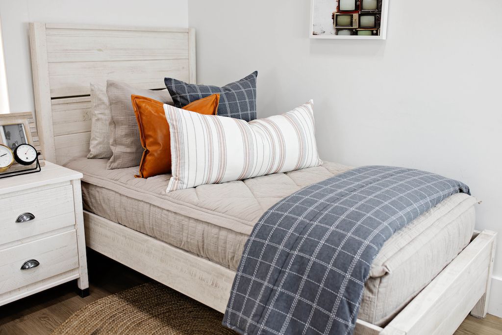 twin bed with tan textured bedding and and blue grid euro, faux leather pillow, cream striped XL lumbar and blue grid blanket at the foot of the bed