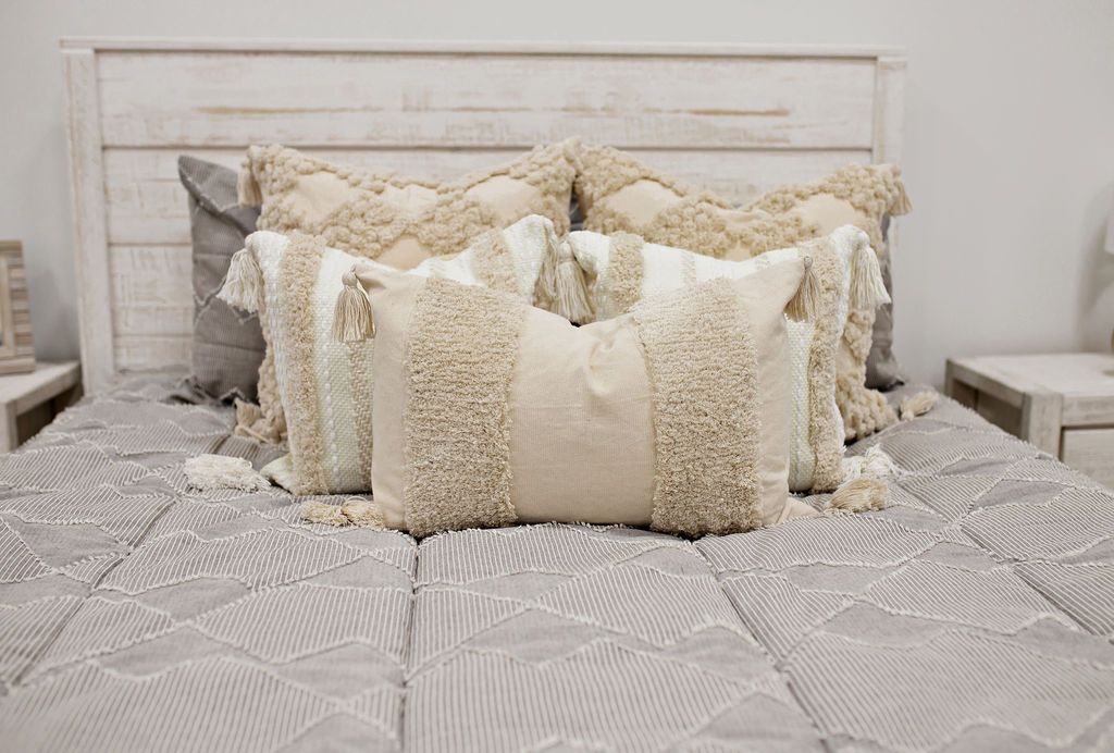 Taupe bedding with textured zig zag design two dark creamy textured euros, two  cream and tan woven textured pillow and a textured dark creamy lumbar with tassels with a cream knitted chenille throw with pom poms along the edge